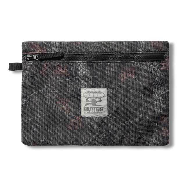 A small zippered pouch with a dark camouflage pattern featuring brown leaf motifs, a black zipper, and a central white square patch with the Butter logo.