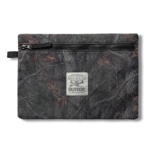 A small zippered pouch with a dark camouflage pattern featuring brown leaf motifs, a black zipper, and a central white square patch with the Butter logo.